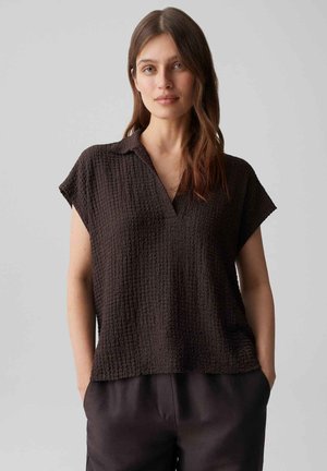 Femme aux longs cheveux bruns portant un haut texturé marron foncé à manches courtes et un pantalon foncé, les mains dans les poches, fond neutre.