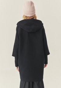 Námořnický oversized kabát s kapucí, který má široké rukávy a žebrované manžety. Nošený s světle růžovou žebrovanou čepicí. Hladká textura, volný design.