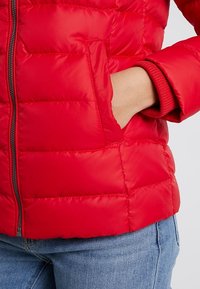 Hånd i forreste lomme på rød quiltet jakke båret med blå jeans, som viser lynlås og ribbet detalje ved manchetterne.