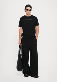 Uomo con capelli pettinati all'indietro che indossa occhiali da sole neri, una maglietta nera, pantaloni larghi neri, mentre tiene una grande borsa a tracolla nera, in piedi su uno sfondo bianco.