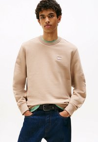 Beige sweatshirt met een ronde hals en geborduurd logo op de borst, gelaagd over een mintgroen t-shirt, gecombineerd met donkerblauwe jeans.