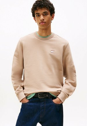 Beige sweatshirt met een ronde hals en geborduurd logo op de borst, gelaagd over een mintgroen t-shirt, gecombineerd met donkerblauwe jeans.