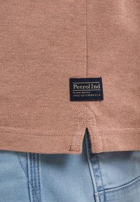 Bavlněné tričko v světle růžové barvě s texturovaným povrchem, na spodní straně s modrým obdélníkovým logem. Světle modré džínové kraťasy viditelné dole.