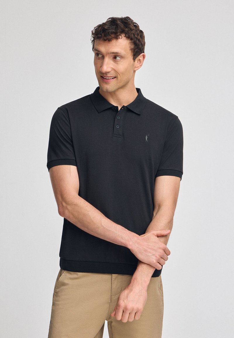 Svart pikétröja gjord av andningsbart tyg med klassisk krage, tre-knappslå och diskret logobroderi på bröstet.