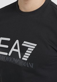 T-shirt nera in cotone con un grande logo bianco "EA7" e il testo "EMPORIO ARMANI" in un accento rettangolare nero sulla parte anteriore.
