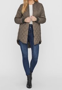 Chaqueta acolchada de color verde oliva con dobladillo redondeado, puños acanalados y cierre de presión en la parte delantera. Lleva puesta sobre una camiseta blanca y unos vaqueros azules.