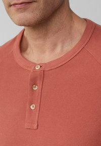 Koszulka henley w kolorze rdzy, wykonana z teksturowanej tkaniny, z prążkowanym kołnierzem, trzema guzikami i kontrastowymi detalami w szwie.