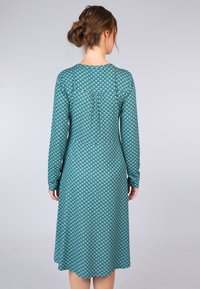Langärmliges Kleid aus teal-farbigem Stoff mit einem Diamantmuster, tailliertem Oberteil und ausgestellt geschnittenem Saum, mit einem Keyhole-Detail am Rücken.