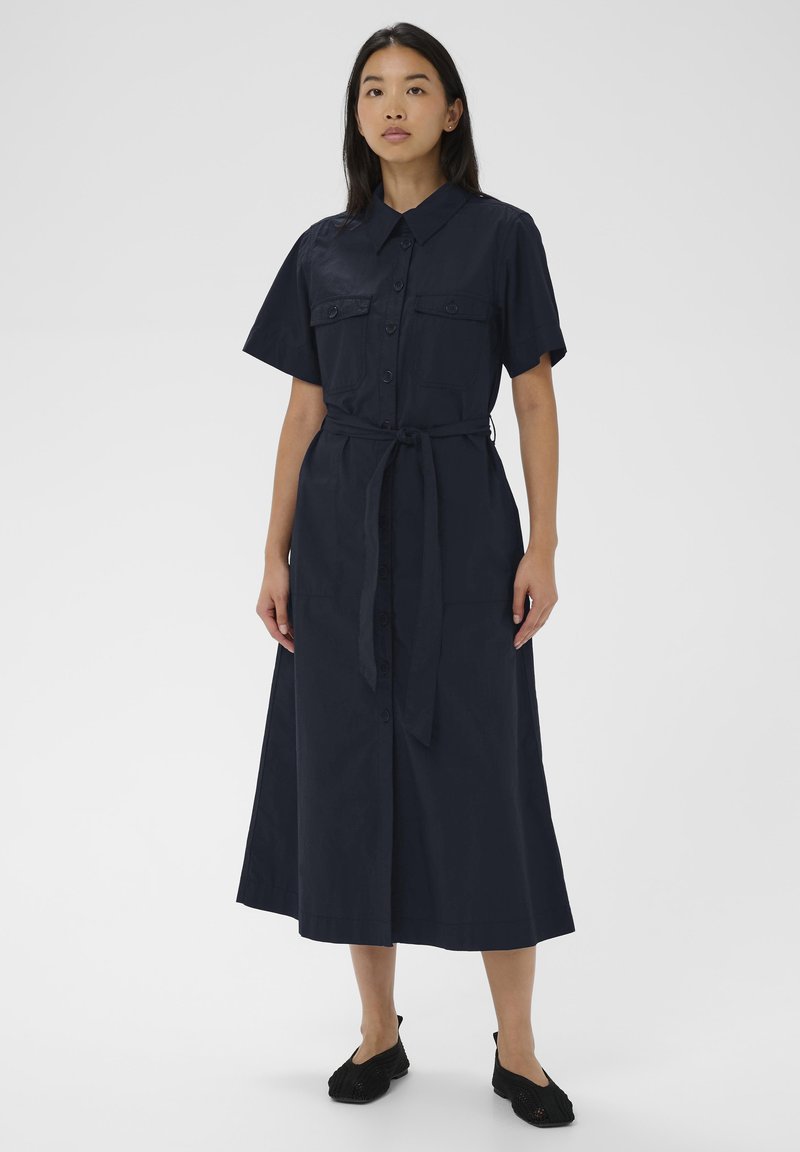 Femme portant une robe midi bleu marine à manches courtes avec boutons sur le devant, ceinture à la taille et chaussures plates noires, debout devant un fond uni.