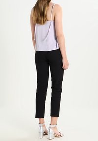 Kvinna med långt hår som bär en ljuslila ärmlös topp, svarta slim fit-byxor och silverfärgade klacksandaler med blockklack, stående vänd bort.