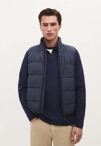 Gilet imbottito blu marino con colletto alto, design a righe orizzontali, chiusura con zipper e tasca frontale; indossato sopra una camicia blu marino a maniche lunghe.