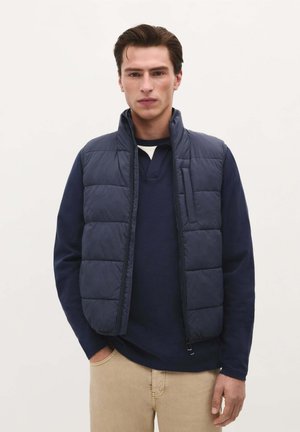 Vesta puffer de culoare navy, cu guler înalt, design cu quiltare orizontală, închidere cu fermoar și un buzunar frontal; purtată peste un tricou cu mânecă lungă de culoare navy.