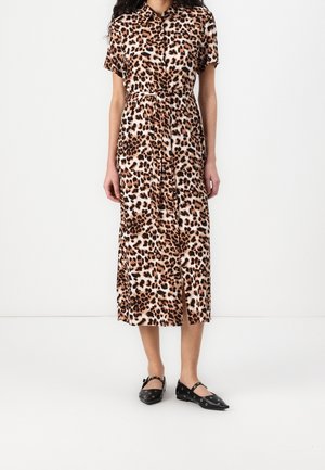 Frau trägt ein knielanges, kurzärmeliges Kleid mit Leopardenmuster, das vorne mit Knöpfen versehen ist, und schwarze flache Schuhe mit Knöchelriemen.