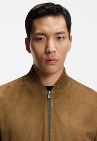 Ung man med kort svart hår, iklädd en brun mockajacka med dragkedja framtill över en svart tröja, vänd framåt med ett neutralt uttryck.