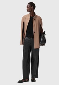 Cappotto a mantella color cammello sopra un maglione nero a trame, abbinato a pantaloni in pelle nera a vita alta e scarpe flat nere, mentre tiene una borsa nera.