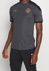 Dark gray athletic shirt with black accents, short sleeves, and Manchester City logo in bronze. Made from moisture-wicking DryCELL material.