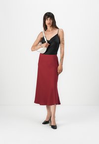 Top en satin noir à fines bretelles, jupe bordeaux mi-longue avec une légère évasement, chaussures noires à talons hauts et un petit sac à main blanc.