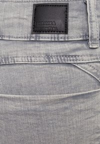 Jeans de mezclilla gris claro con una etiqueta de cuero que dice "BERSHKA ESSENTIAL", con detalles de costura y diseño estándar de cinco bolsillos.