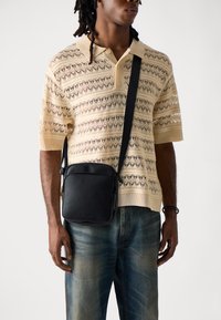 Black leather crossbody bag with a smooth texture, square shape, and a front zip closure, worn over a light knit polo shirt and blue jeans.