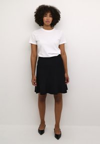 Svart utsvängd kjol i mjukt tyg, kombinerad med en vit t-shirt med kort ärm. Modellen bär svarta spetsiga klackskor och står mot en neutral bakgrund.