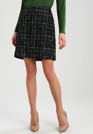 Vrouw draagt een zwarte geruite rok tot de knie, een groene top met lange mouwen en beige hoge hakken, staand tegen een effen lichte achtergrond.
