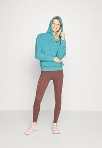 Sweatshirt com capuz e cordões, combinado com leggings castanhas e sapatilhas atléticas de rosa claro. Modelo está a sorrir e a posar.
