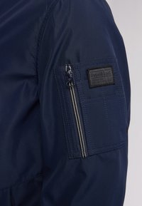 Marineblauwe waterdichte jas met een ritspocket op de bovenarm, voorzien van een gestructureerd zwart label met "Petrol Ind" merknaam.