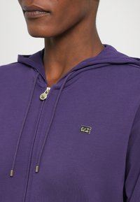 Paars zip-up hoodie gemaakt van zachte stof, met een klein gouden logo, een trekkoordkap en een gladde textuur, gezien van hals omhoog.