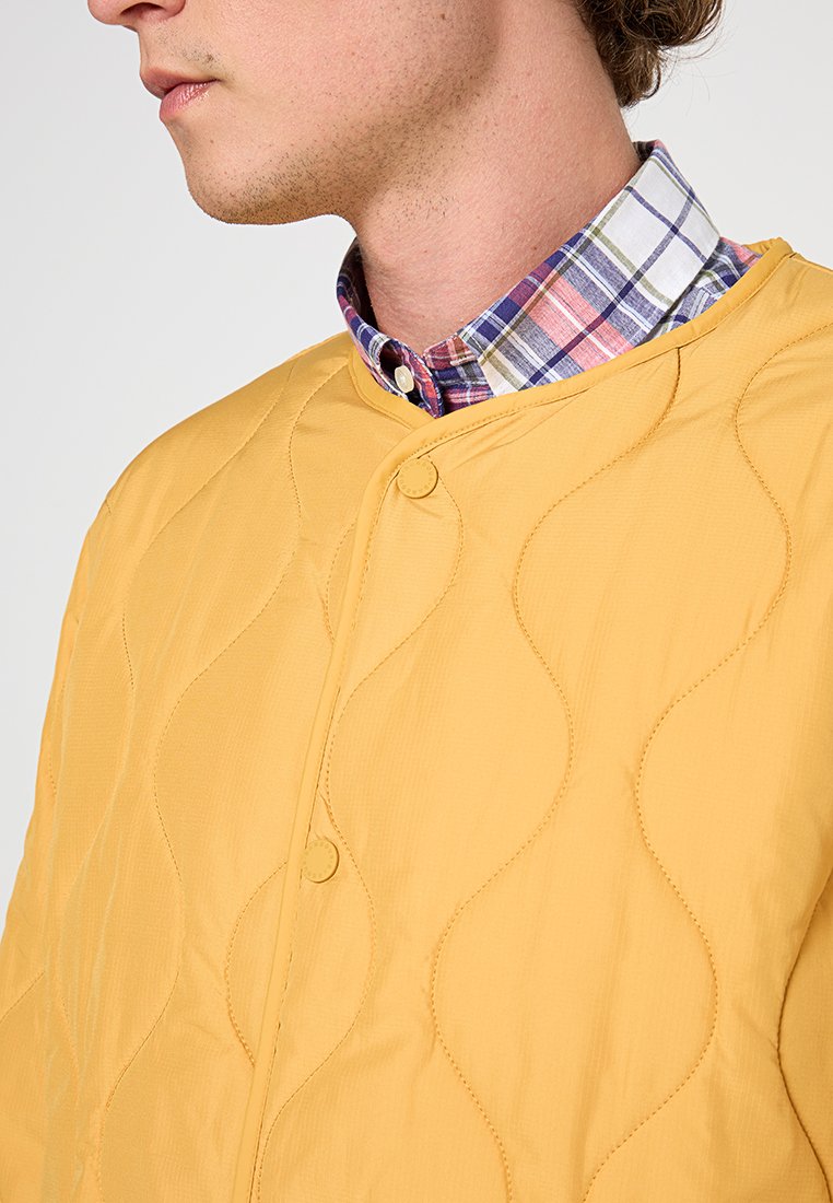 Mann in einer gelben gesteppte Jacke über einem mehrfarbigen karierten Hemd, sichtbar von den Schultern aufwärts, nach links schauend.