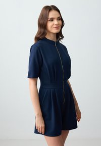 Marineblauwe short-sleeve jumpsuit met een hoge halslijn en gouden ritsdetails. Heeft een getailleerde taille en geplooide short. Van een katoenmix materiaal.
