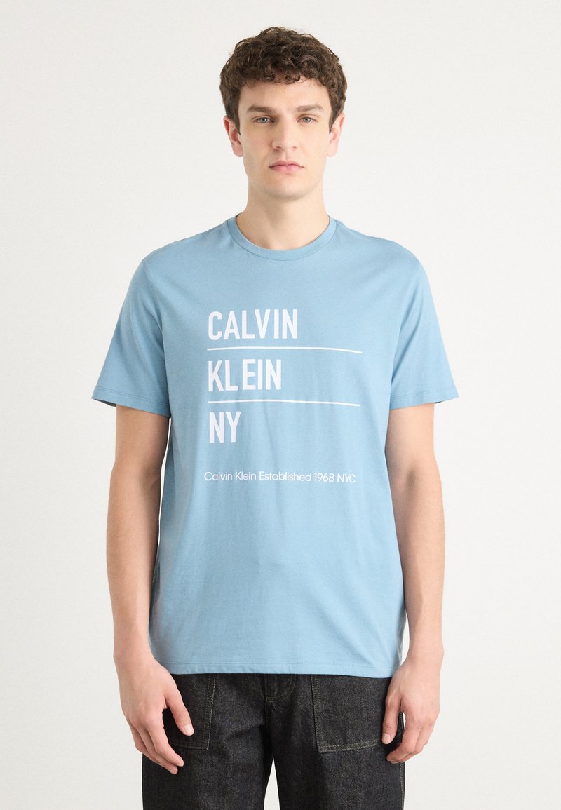Šviesiai mėlynos medvilnės marškinėliai su baltu užrašu "CALVIN KLEIN NY" ir "Calvin Klein Established 1968 NYC." Trumpomis rankovėmis ir apvaliu iškirpimu.