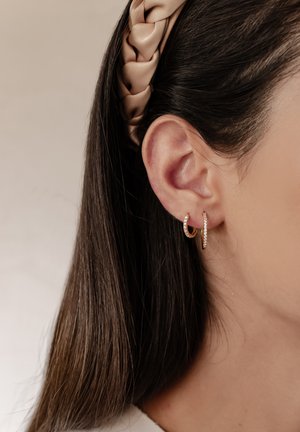 Gold hoop earrings with small clear stones, worn on a model's ear. Hair partially tucked behind a satin cream-colored headband.