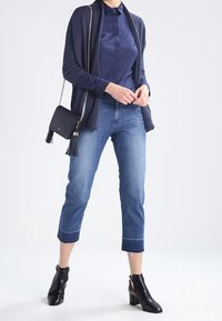 Marinblå cardigan, tajt skjorta och korta blå jeans. Svarta klackstövlar vid vristen. Liten svart väska med fransdetalj. Släta texturer och moderna linjer.