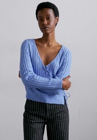 Pulover ribbed de un albastru deschis, cu mâneci lungi, decolteu adânc în V și detalii cu legături pe laterale, asociat cu pantaloni negri cu dungi subțiri. Textură moale.