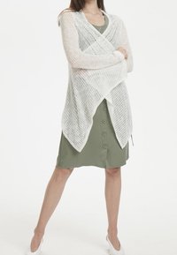 Bílý otevřený pletený cardigan přetažený přes zelené šaty s knoflíky. Cardigan má volnou, texturovanou vazbu a splývavý design.