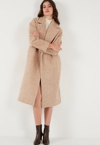 Manteau en mélange de laine texturé beige avec un design à double boutonnage, des revers larges et une coupe mi-longue, doté d'une fente à l'avant et de poches latérales.
