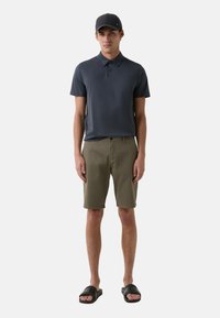 Pánský outfit složený z tmavě modrého polokošile, olivových šortek a černých nazouvacích sandálů. Polokošile má límec a jemný detail loga.
