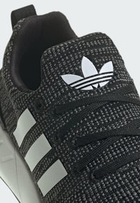 Scarpa sportiva nera con tomaia in maglia testurizzata, caratterizzata dal logo a tre strisce bianche, lacci e un accento bianco con il marchio sulla linguetta.