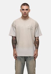 Beżowy, oversize t-shirt z krótkimi rękawami, z delikatnym logo nadrukowanym na klatce piersiowej. Materiał wydaje się miękki, o gładkiej teksturze.