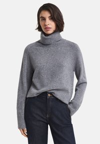 Donna con un maglione grigio a collo alto infilato in jeans blu scuro, in piedi con una mano in tasca su uno sfondo semplice.