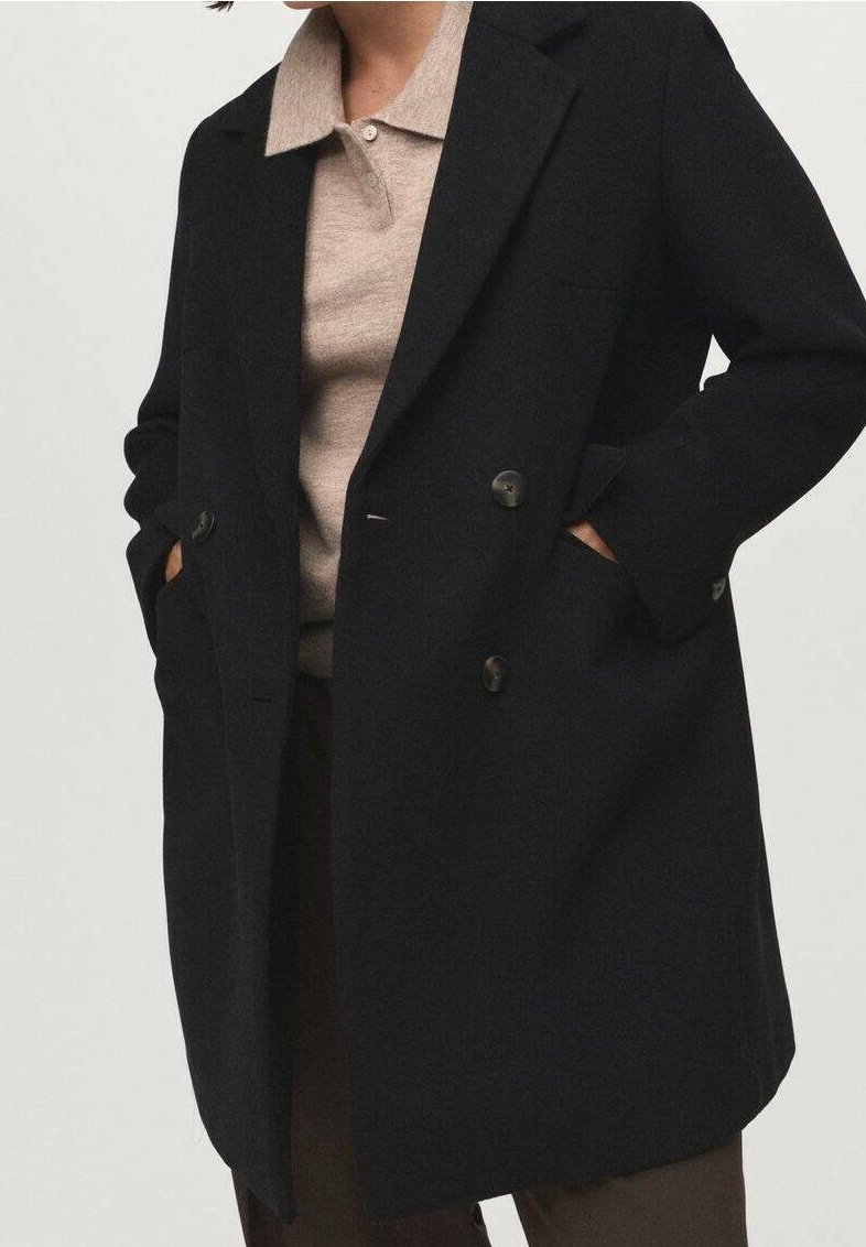 Personne portant un manteau noir croisé sur un pull beige à col, les mains dans les poches du manteau, sur un fond neutre.