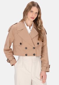 Beskuren beige trenchcoat med dubbelknäppt knappar, vida slag och justerbara ärmslut med bälte, kombinerad med en vit skjorta och ljusa byxor.