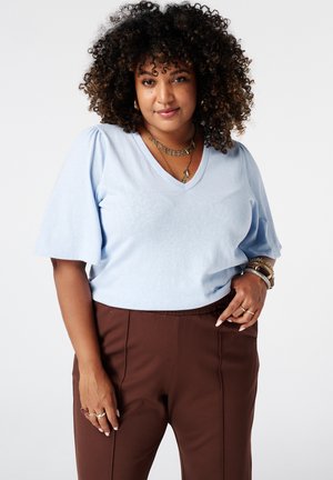 Vrouw met krullend haar die een lichtblauwe V-hals blouse en een bruine broek draagt, geaccentueerd met armbanden en gelaagde kettingen, staand tegen een witte achtergrond.
