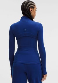 Haut de sport à manches longues en bleu foncé avec une coupe ajustée, tissu lisse, col montant et détails avec couture au dos. Logo visible à l'arrière.