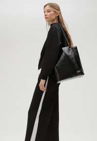 Černá kožená tote taška s texturovaným povrchem, vyznačující se zlatým kováním a minimalistickým designem, kterou nese osoba oblečená v černém.