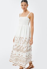 Abito maxi in cotone bianco con corpetto arricciato, spalline sottili e intricati ricami marroni lungo l'orlo che raffigurano motivi floreali e geometrici.