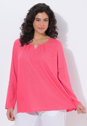 Kobieta o kręconych, ciemnych włosach, ubrana w jasnoróżową bluzkę z długim rękawem, trzymająca rant bluzki, stojąca na tle jednolitego białego tła.