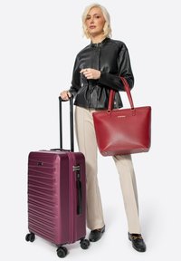 Valigia rigida viola con texture a righe e manico retrattile, accanto a una borsa tote rossa in pelle con fondo piatto e cuciture decorative.