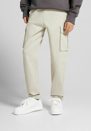 Pantalon cargo beige avec poches à rabat latérales porté avec des baskets blanches et un haut à manches longues gris foncé, montré de la taille aux pieds.