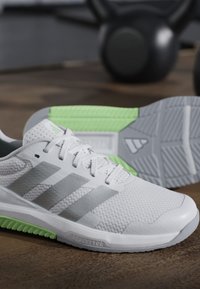 Chaussure de sport blanche et grise avec des accents verts sur la semelle, posée sur un sol en bois avec des kettlebells noirs flous en arrière-plan.